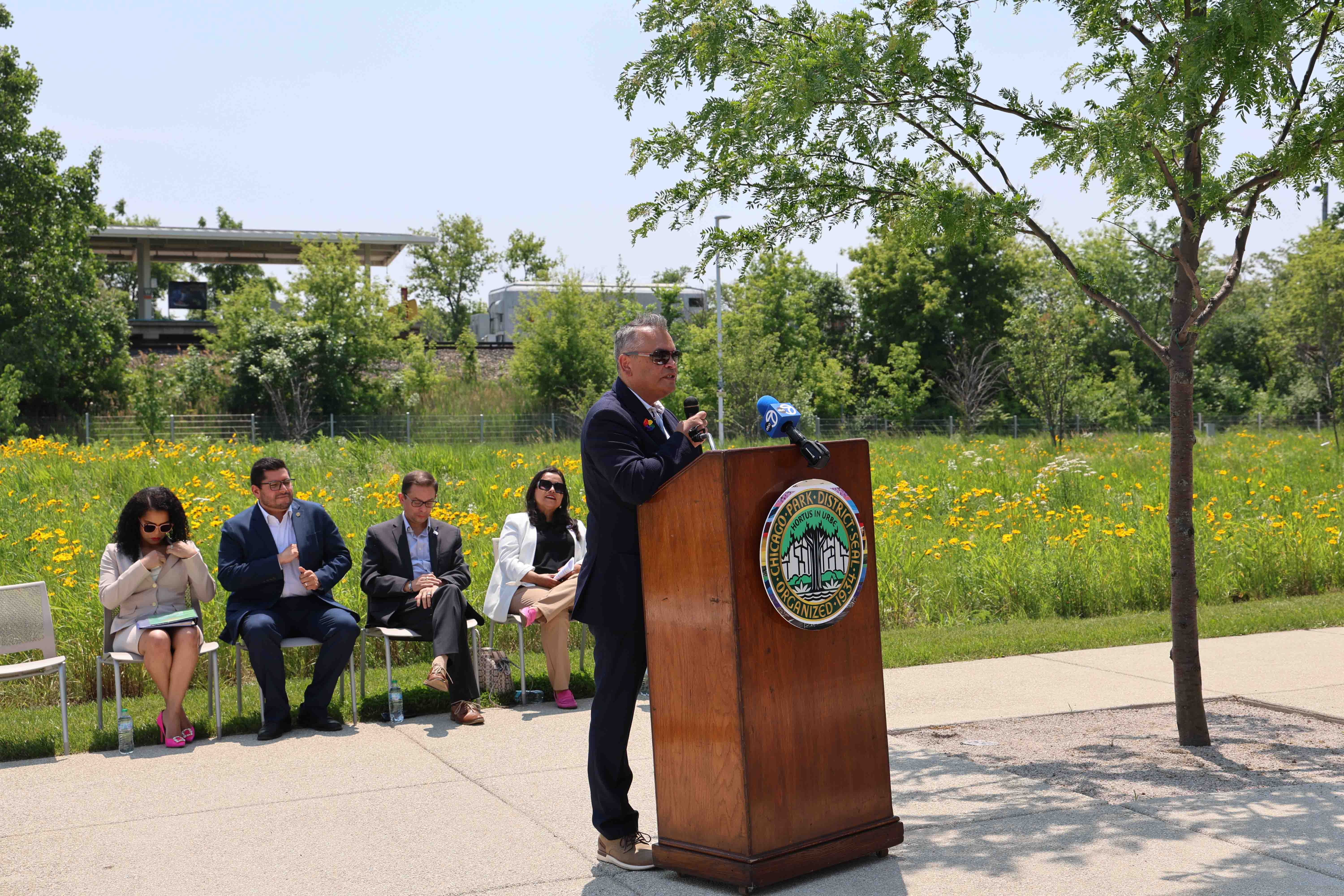 Man speaks at podium with Chicago Park District seal, audience seated nearby. Train station and wildflowers in background.